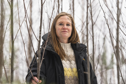 Person i vinterkläder som står bland täta, bladlösa stammar i en snöklädd ungskog.