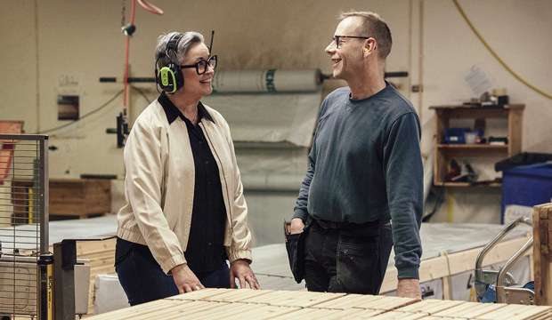 Två personer i en fabriksmiljö, en med ljus jacka och hörselskydd, den andra med mörk tröja och händerna på höfterna, omgivna av trämaterial och utrustning.