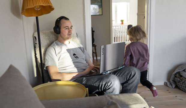 Person sitter i en fåtölj med hörlurar och arbetar vid en bärbar dator i ett hem, medan ett barn rör sig i rummet intill.